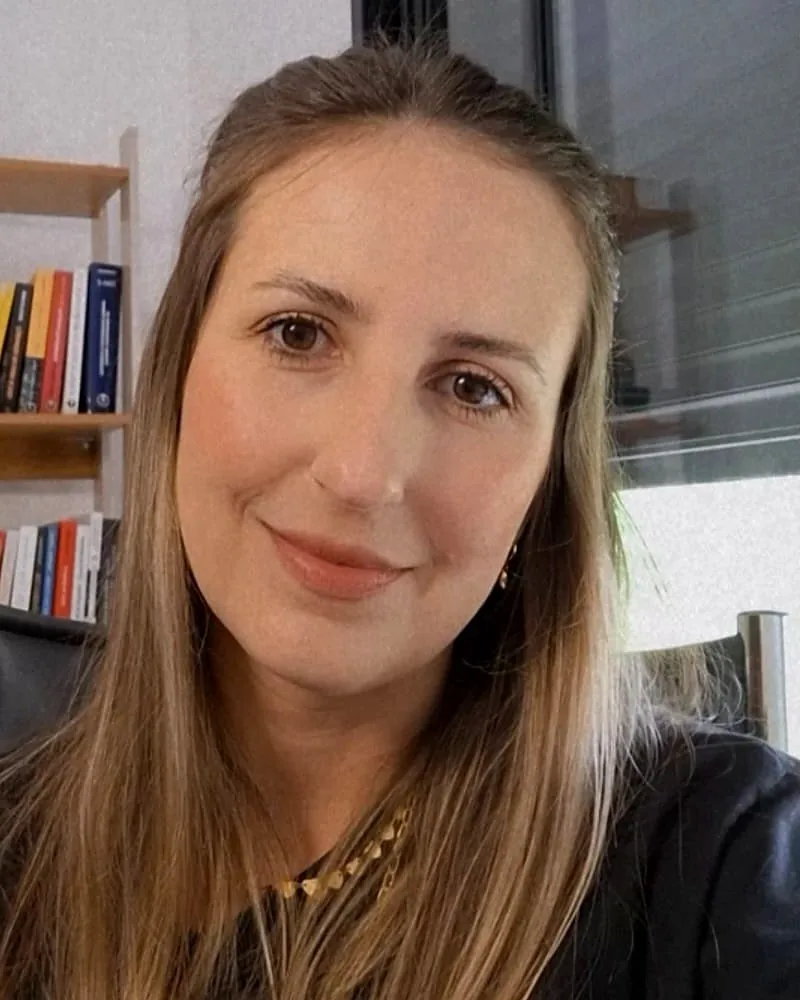 Mariana Hermann, psicóloga, sorrindo em seu consultório, com uma estante de livros ao fundo.